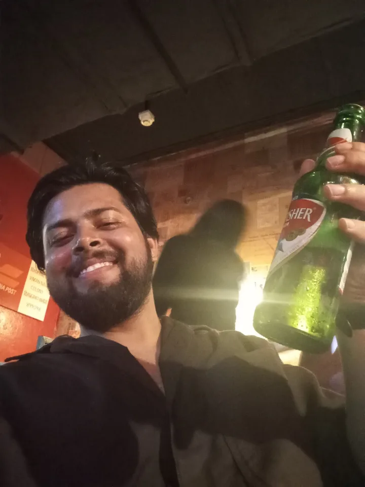 Yoginder smiling at the camera with a bottle of Kingfisher beer held up, in a dim bar
